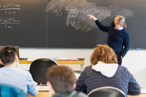 A faculty member, wearing a mask, writes on a chalkboard.