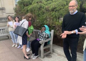 President Nicole Farmer Hurd stands before Fatimata Cham, who is sitting on a bench, with flowers. Others around them clap and smile