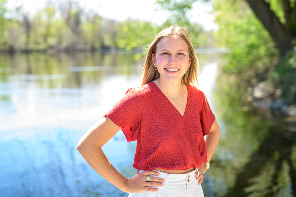 Libby Mayer smiles with her hands on her hips, outside of a scenic river.