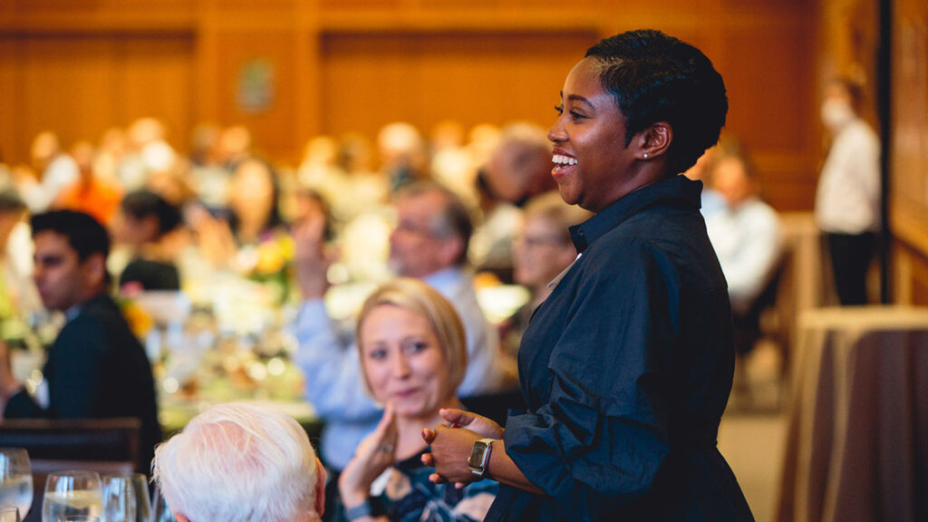 Kamaka Martin stands in a room where College faculty and staff sit an applaud her