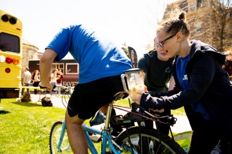 student in a blue T-shirt rides on a stationary bike on the Quad, students hold a blender container behind him