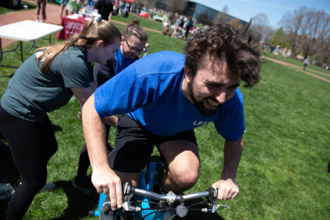 student in a blue T-shirt rides on a stationary bike on the Quad, students hold a blender container behind him