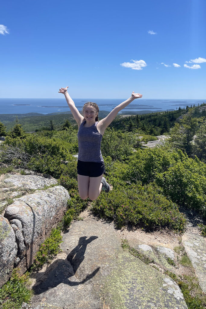 Lucy Kade '22 jumps in the air with arms up, in front of a scenic view.