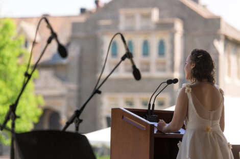 student in white dress speaks at podium, Hogg in background