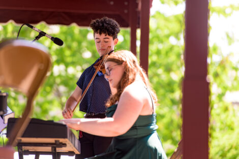 student plays violin, while another student plays keyboard, green trees in background