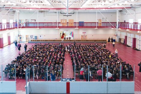 graduates in caps and gowns sit in chairs arranged in rows in gym