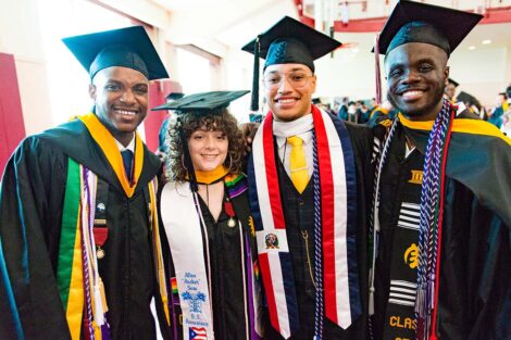four students wearing caps, gowns, honor cords stand together and smile in gym
