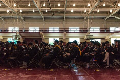 graduates in caps and gowns sit in chairs arranged in rows in gym