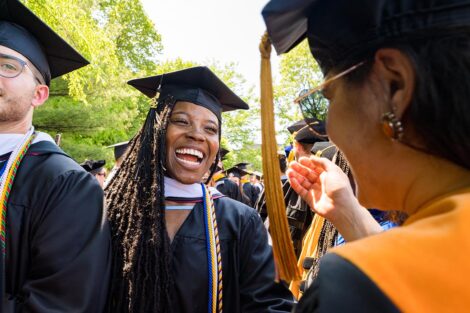 student in cap and gown smiles and laughs as loved one reaches out to embrace her