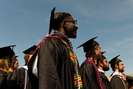 students in cap and gowns stand and smile, blue sky behind them