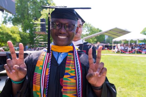 student holds up hands giving two peace signs while smiling and wearing cap and gown