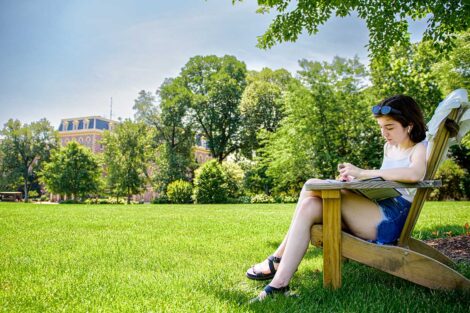 student sits on an Adirondack chair on the Quad, green grass is in front of her, Pardee is in the background