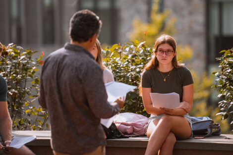 A professor holds class outside, speaking to their class surrounded by trees.