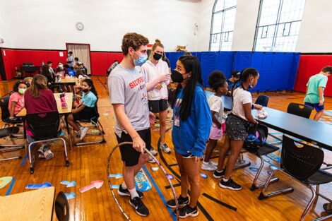 Lafayette students wearing masks stand in a gymnasium where young children are gathered doing activities at tables
