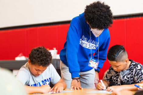 a Lafayette student wearing a mask leans over a table where two young children color with markers