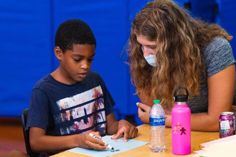 a Lafayette student in a mask sits at a table with a young student who is coloring with a marker