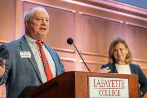 Michael Weisburger is standing at a podium speaking to the audience. He is wearing a light blue jacket and a red tie.