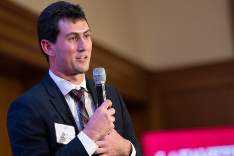 Joshua Wolf is wearing a black jacket and a maroon striped tie and is holding a microphone.