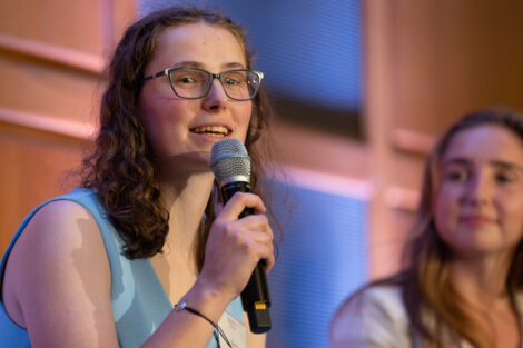 Becca is wearing a light blue sleeveless dress and is holding a microphone.