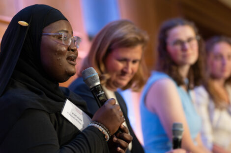 Fatimata Cham is holding a microphone and speaks to the audience. She is wearing a black head scarf and a black shirt.