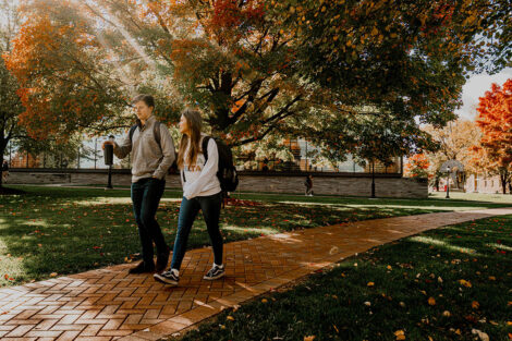 Two students walk along a brick path surrounded by trees with colorful leaves.