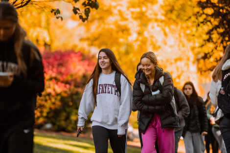 Two students walk along a brick path surrounded by trees with colorful leaves.