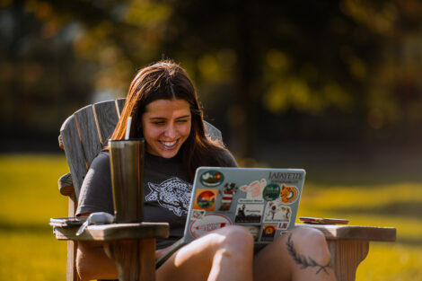 A student smiles, sitting outdoors with their laptop